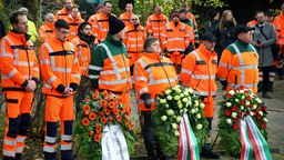 Mehrere Straßenwärter in orangefarbenen Uniformen stehen vor Kränzen