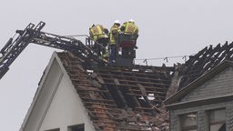 Drei Feuerwehrleute stehen auf einem Kran beim Dachstuhl des ausgebrannten Hauses. 