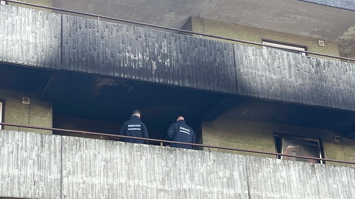 Auf dem Bild sind die Brandspuren der Wohnung außerhalb des Gebäudes zu sehen. Zwei Polizisten stehen auf dem Geländer.