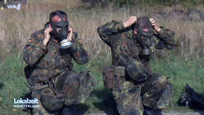 Zwei Menschen in Bundeswehruniform setzen Gasmasken auf.