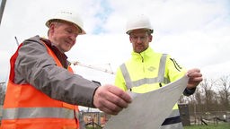 Zwei Männer in Warnkleidung und mit Helm betrachten Plan auf Baustelle