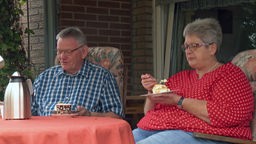 Ein älterer Mann in einem blau-weiß karierten Hemd und eine ältere Frau in roter Bluse essen Kuchen auf einer Terrasse. 