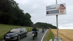 Ein schwarzer VW Golf und Motorräder auf einer Landstraße, am Straßenrand ein Schild mit der Aufschrift "pssst...RÜCKSICHT!".