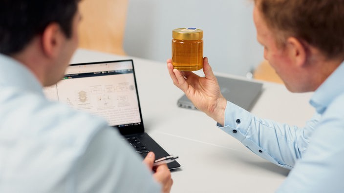 Zwei Männer sitzen vor einem Laptop und halten ein Glas Honig hoch. 