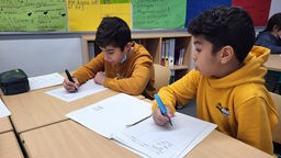 Zwei Jungs schreiben auf Blättern in einem Klassenraum. 