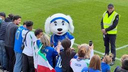 Ein Maskottchen mit Italientrikot steht auf einem Fußballplatz, daneben eine Reihe Zuschauer ebenfalls mit Italientrikots.