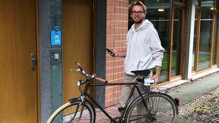 Felix Erdmann steht neben einem Fahrrad und hält den Sensor fest, in der anderen Hand sein Smartphone