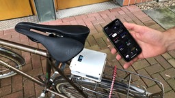 Ein Fahrrad von oben, unter dem Sattel ein Sensor, jemand hält ein Smartphone ins Bild 