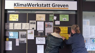 Zwei Menschen hängen Zettel an eine Wand mit dem Titel KlimaWerkstatt Greven. 
