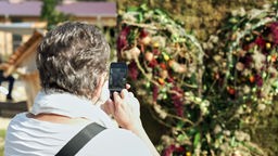 Eine ältere Frau von hinten, die mit ihrem Smartphone eine Blumen-Gesteck in einem Park fotografiert