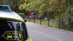 Ein Krankenwagen steht hinter einem Zaun, im Vordergrund ist ein Teil eines Polizeiautos zu sehen