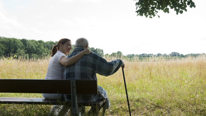 Ein Senior mit Stock und eine Pflegerin sitzen auf einer Bank an einer Wiese