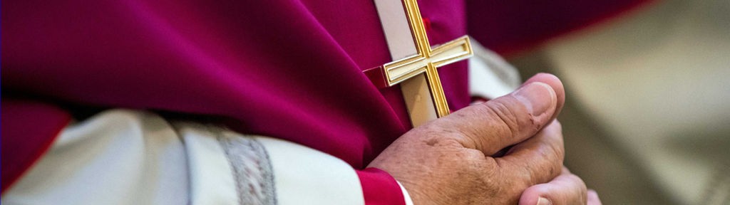 Die gefalteten Hände eines Bischofs im Gewand. Der Bischof trägt eine Kette mit einem goldenen Kreuz.