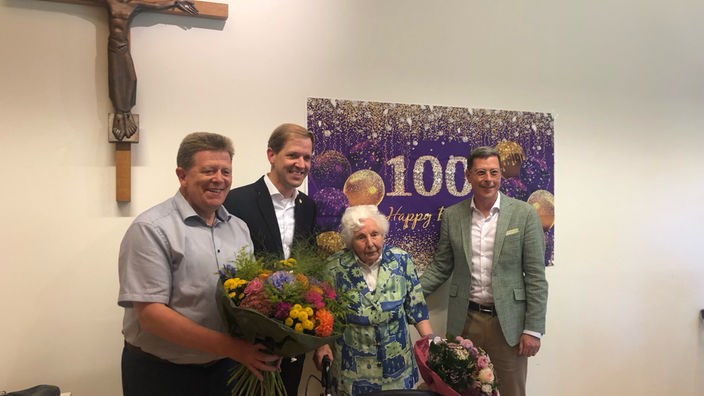 ältere Frau steht mit Rollator vor einem Schild auf dem 100 udn Happy Birthday steht. Sie ist umgeben von drei Männern, die zum Geburstag gratulieren.