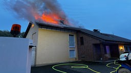 Man sieht ein Einfamilienhaus, aus dem Dach steigen Flammen. Vor dem Haus ist ein Feuerwehrmann.