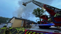 Man sieht ein Einfamilienhaus, aus dessen Dach steigen Flammen. Davor stehen Feuerwehrmänner, sowie ein Feuerwehrauto und ein Rettungskran.