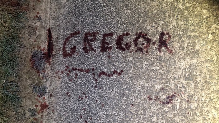 Ein asphaltierter Weg, auf dem mit Blut in Großbuchstaben "Gregor" steht. 