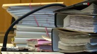 Mehrere dicke Ordner mit Papier liegen in einem Gerichtssaal