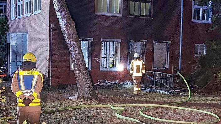 Ein Haus von außen mit zwei Feuerwehrleuten davor und einer ausgebrannten Erdgeschosswohnung