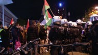 Polizisten mit Helmen stehen vor Protestanten, die Palästina-Flaggen halten.