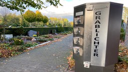 Ein Automat auf dem Grablichter steht, dahinter ein Friedhof