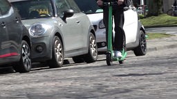 Eine Person auf einem E-Scooter fährt auf einer Straße an parkenden Autos vorbei.