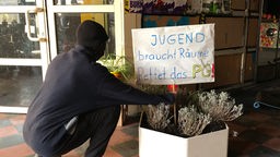 Eine vermummte Person steckt ein Schild in einen großen Blumentopf. Die Aufschrift lautet: "Jugend braucht Räume. Rettet das PG!"