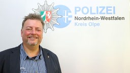 Polizist Michael Meinerzhagen lächelt in die Kamera, im Hintergrund der Schriftzug der Polizei Olpe mit Logo