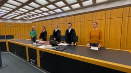 Im Gerichtssaal, fünf Personen hinter einem Tisch stehend.