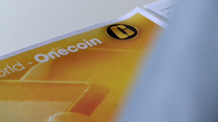 Auf einem weißen Tisch liegt ein Blatt Papier darauf gedruckt das Onecoin-Logo.