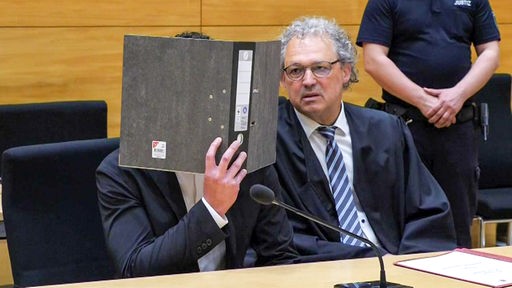 Ein Mann sitzt auf der Anklagebank mit einem Ordner vor dem Gesicht, neben ihm sein Anwalt. 