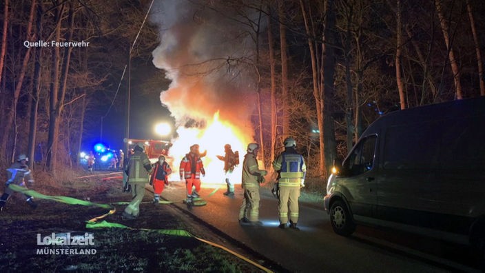 Brennendes Auto  in einer Waldstraße, mit Feuerwehrmännern im Einsatz. 
