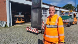 Mann steht in orangefarbener Arbeitskleidung vor den offenen Toren eines Fahrzeuglagers der Autobahnmeisterei.