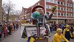 Die Figur "Gru" von den "Minions" auf einem kleinen Karnevalswagen beim Münsteraner Rosenmontagsumzug, am Straßenrand Karnevalisten und Karnevalistinnen.