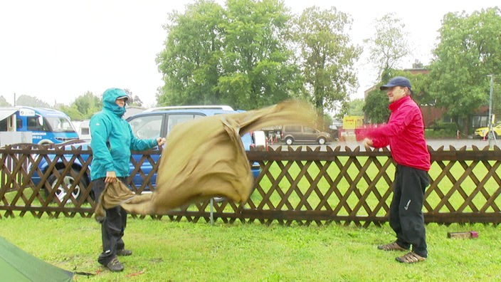 Auf dem Foto sind eine Frau und ein Mann in Regenjacken, die zu zweit eine Plane schütteln, um sie von Regen zu befreien.