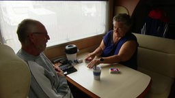 Auf dem Foto sind ein Mann und eine Frau, die bei Regen in einem Wohnmobil an einem Tisch Karten spielen.