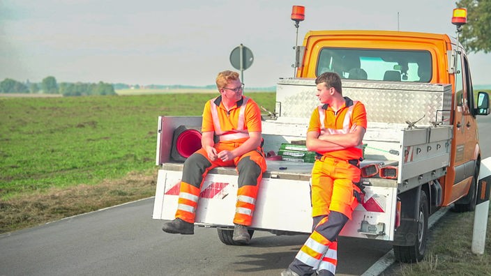 Zwei junge Männer in Arbeitskleidung sitzen an einem Auto.