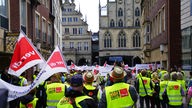 Auf dem Foto sind Menschen in gelben Warnwesten, die mit Verdi-Flaggen durch die Innenstadt Münsters ziehen.