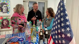 Drei Menschen an einem Stehtisch mit Getränkeflaschen und US-amerikanischer Flagge