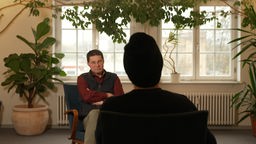 Im Hintergrund zu sehen ist Gestalttherapeut Helge Rettig, im Vordergrund, mit dem Rücken zur Kamera, sitzt Lukas