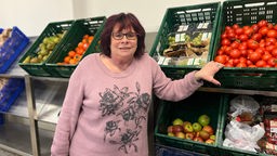Eine Frau mit rötlichen Haaren, Brille und rosa Strickpulli steht vor Gemüsekisten 