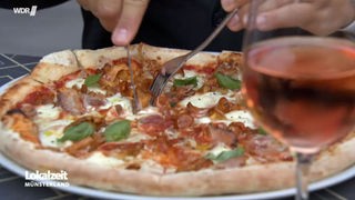 Eine Pizza mit Pilzen und Spanferkel, die gerade angeschnitten wird, im Vordergrund ein Glas Wein 