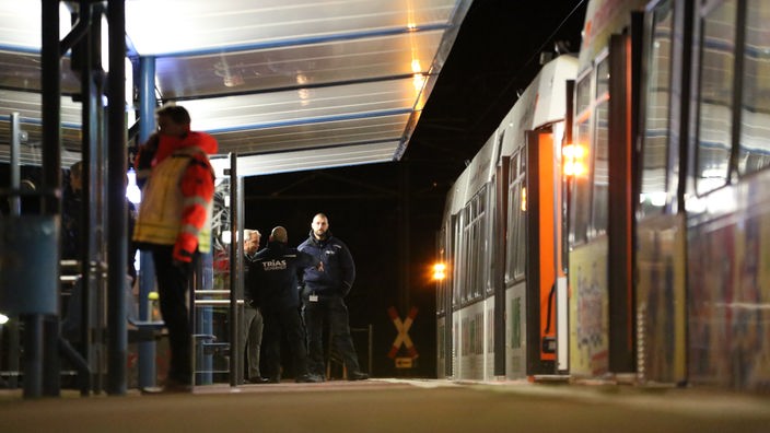 An einer Stadtbahnhaltestelle in Bielefeld bei Nacht telefoniert eine Rettungskraft. Im Hintergrund stehen einige Sicherheitskräfte. Rechts steht die Stadtbahn.