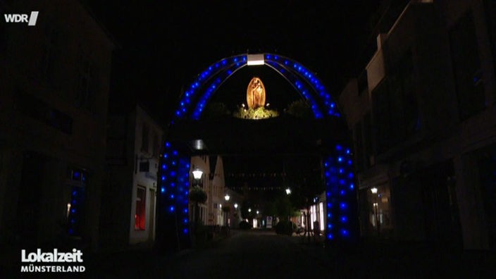 Ein blau leuchtender Bogen bei Nacht über einer erleuchteten Marienstatue.