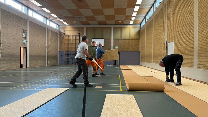 Vier Männer verlegen Holzplatten auf dem Boden in einer Turnhalle.