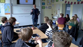 Ein Lehrer steht mit Laptop in der Hand vor der Schulklasse