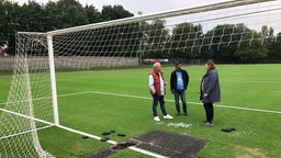 Zwei Männer und eine Frau stehen auf einem Fußballplatz vor einem Fußballtor, das zuvor zerschnitten wurde. Vor ihnen fehlt auch ein rechteckiges Stück des Rasens