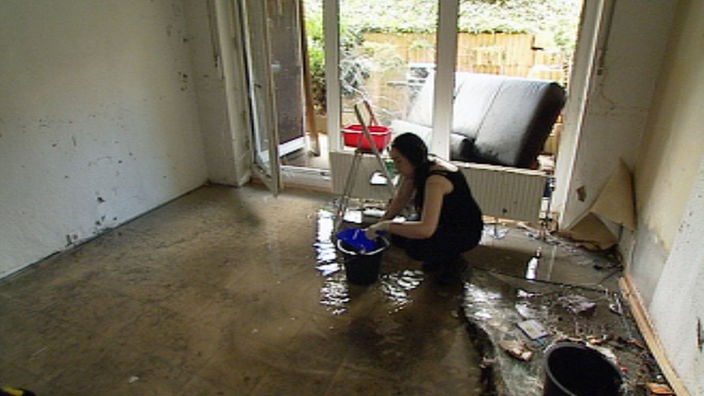 Frau kniet vor Wischeimer in zerstörtem Wohnzimmer