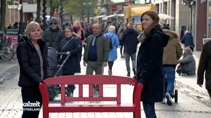 Zwei Frauen mit einer roten Bank stehen in der Fußgängerzone in der Ludgeristraße in Münster. 