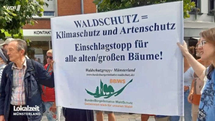 Zwei Personen auf einer Demonstration halten ein Schild für "Waldschutz im Münsterland" in die Höhe.